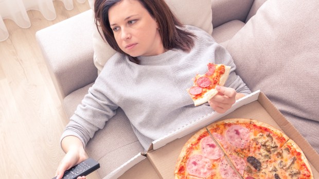 woman eating pizza and holding phone laying on sofa at home