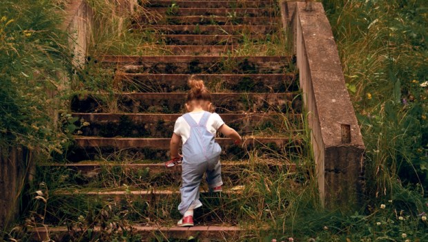 A little girl, 2 years old, with a ponytail and in a jumpsuit, climbing up the steps of the old stone