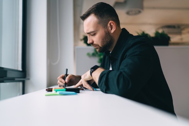 Side view of concentrated male influencer writing down information in sticky note with pen while working in office sitting in front of window | career,workspace,occupation,focus,project,office,employee,professional,horizontal,side view,startup,casual,pen,sticky note,businessman,plan,write,beard,work,take note,specialist,marker,job,note,young,idea,smart,influencer,modern,busy,indoors,company,man,table,sit,business,manager,sticker,staff,concentrate,entrepreneur,intelligent,marketing,corporate,hardworking,analyst,information,strategy,workplace - shutterstock_2336815877.jpg