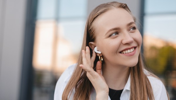 Happy blonde woman sitting on the bench near office building with wireless earbuds in ears